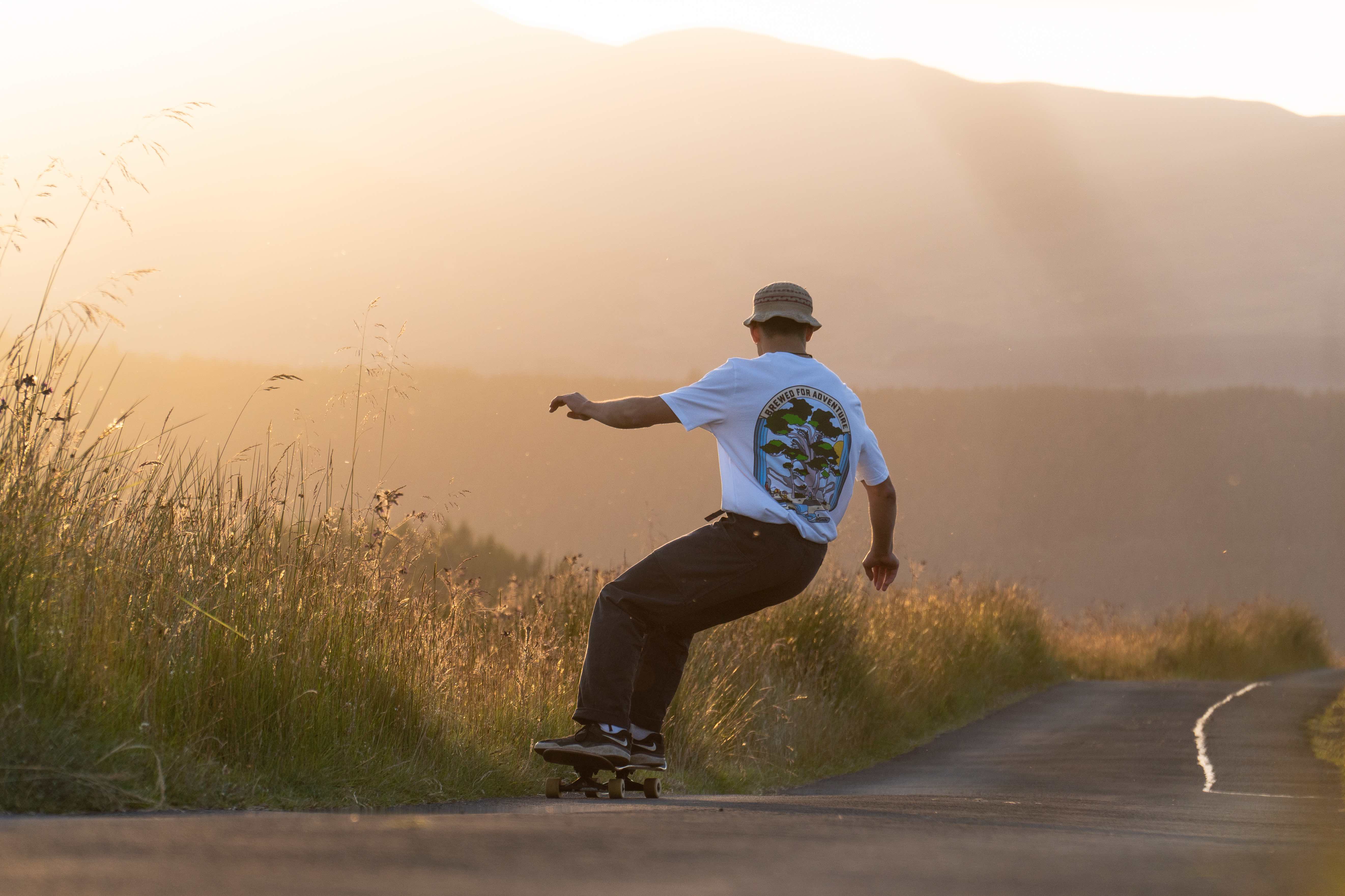 A person skateboarding down a country road wearing a white Brewed for Adventure Glen Lyon T-shirt